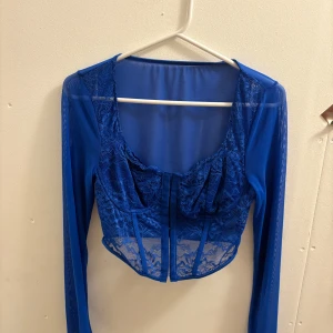 Blå mesh korsettopp med spetsdetaljer  - Snygg blå korsettopp i mesh med spetsdetaljer och långärmade ärmar. Toppen är croppad och har en markerad byst med transparent tyg och dekorativa sömmar framtill. Perfekt för dig som vill sticka ut med en trendig och edgy look.