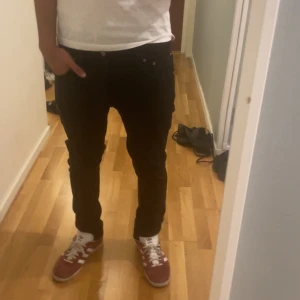 Svarta jeans från Nudie Jeans - Säljer ett par svarta jeans från Nudie Jeans med klassiska bakfickor och diskret logga. Jeansen har en rak passform. Inga defekter. Storleken är 30/32  och personen på bilden är 180 cm samt väger 70 kg
