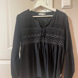 Svart blus med broderi och knyt - Supersnygg svart blus med broderade detaljer och knyt framtill. Långärmad modell med smock och diskret mönster över bröstet samt broderad detalj på ryggen. Luftig passform i mjukt bomullstyg, perfekt till jeans eller kjol.