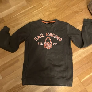 Mörkgrön Sail Racing sweatshirt - Snygg mörkgrön sweatshirt från Sail Racing med tryck i vitt och orange framtill. Tröjan har rund halsringning, långa ärmar och mjukt bomullsmaterial. Perfekt för dig som gillar sportig och avslappnad stil.