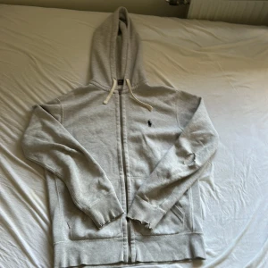 Grå hoodie från Polo Ralph Lauren - Grå hoodie med dragkedja från Polo Ralph Lauren, klassisk design med huva och snörning. Tröjan har långa ärmar, ribbade muddar och fickor framtill. Ikonisk liten Polo-logga broderad på bröstet. Perfekt för chill dagar och streetwear-vibe. Den har et litet hål på höger som knappt syns och ett större på vänster och på arm enden så är det också hål på båda därför säljs den så billigt nypris 2229kr
