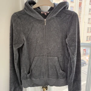 Grå Juicy Couture hoodie med strass - Säljer en grå hoodie från Juicy Couture i storlek S. Tröjan har huva, dragkedja med J-detalj och fickor fram. På ryggen finns ett rosa strassmönster med Juicy Couture-logga. Materialet är mjukt och känns lyxigt, perfekt för chill dagar.