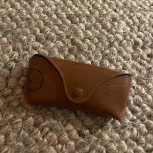 Ray-Ban brun glasögonfodral - Snyggt brunt glasögonfodral från Ray-Ban i klassisk design. Fodralet är gjort i ett läderliknande material med tryckt logga på framsidan och knappstängning. Perfekt för att skydda dina solglasögon eller vanliga glasögon.