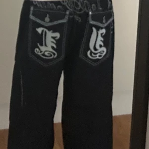 Wide svarta jeans med broderade fickor - Säljer ett par svarta wide jeans från Le Jean de Marithé + François Girbaud med stora broderade fickor bak och kontrastsydda sömmar. Jeansen har coola detaljer med vit brodyr och knappar på ficklocken. Perfekt för dig som gillar streetwear och unik design.