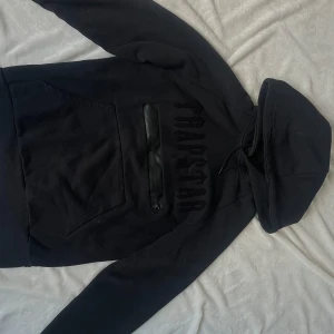 Svart Trapstar hoodie med ficka - Svart Trapstar hoodie med tryck på bröstet och på luvan. Den har även en ficka med dragkedja på magen. 