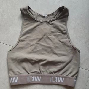 Beige linnetopp från ICANIWILL - Snygg beige linnetopp från ICANIWILL med bred resår nertill där loggan ICANIWILL syns i vitt. Modellen är ärmlös med hög halsringning och brottarrygg. Perfekt för träning eller chill dagar. Mjuk bomullskänsla och stilren design.