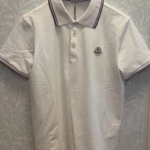 Vit Moncler pikétröja med ränder - Vit pikétröja från Moncler med korta ärmar och klassisk krage. Kragen och ärmsluten har snygga ränder i rött och marinblått. Tröjan har knappar framtill och Moncler-logga broderad på bröstet. Tillverkad i mjuk bomull som känns skön mot huden. Pris kan diskuteras 