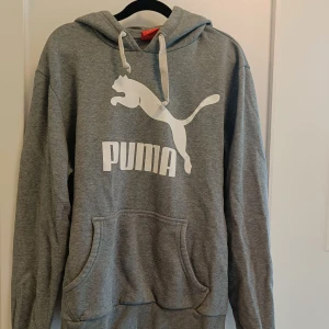 Grå hoodie från Puma med logga - Säljer en grå hoodie från Puma med stor vit logga och text på bröstet. Tröjan har huva med snörning, magficka och ribbade muddar. Perfekt för chill dagar eller när du vill ha en sportig look. Materialet är mjuk bomull och polyesterblandning.