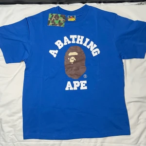 Bape T-shirt blå  - Snygg blå t-shirt från BAPE med stor vit text och klassisk brun ape-logga på bröstet. Rund hals och korta ärmar, tillverkad i mjuk bomull. Taggen har camo-mönster och BAPE-etikett i nacken. Perfekt för dig som gillar streetwear och vill sticka ut.