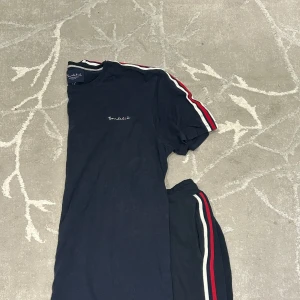 Mörkblå set - Snygg mörkblå t-shirt från Bordéus med broderad logga på bröstet. T-shirten har korta ärmar och coola ränder i rött och vitt längs axlarna. Perfekt för dig som gillar sportig och stilren look.