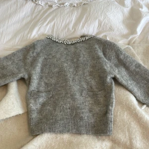 Grå stickad tröja med detalj  - Mysig grå stickad tröja i mysigt material. Tröjan har två små fickor framtill och en snygg pärlbesatt krage som ger extra bling. Perfekt för dig som gillar att sticka ut lite och vill ha något mjukt och fluffigt i garderoben🩷 aldrig använd!