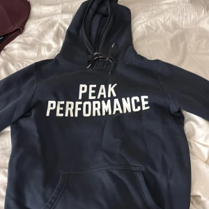 Mörkblå hoodie från Peak Performance - Säljer en mörkblå hoodie från Peak Performance i storlek S. Tröjan har vit text på bröstet, huva med snörning och en klassisk känguruficka framtill. Perfekt för chill dagar och sportiga outfits. Materialet är mjuk bomull och polyesterblandning.