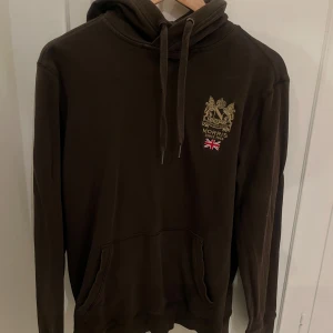 Brun hoodie från Morris med tryck - Snygg olivgrön hoodie från Morris med guldfärgat emblem och engelsk flagga broderat på bröstet. Tröjan har huva med snörning, känguruficka och långa ärmar. Använd men fint skick! 