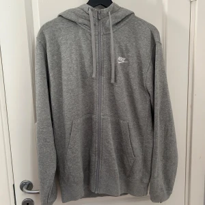 Grå zipper hoodie från Nike - Säljer en grå hoodie från Nike med vit logga på bröstet. Tröjan har huva med snörning, två fickor fram och hel dragkedja. Mjuk insida i fleece och långärmad passform. Perfekt för chill eller träning, enkel att matcha med jeans eller joggers. Priset är förhandlingsbart!