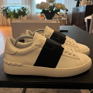 Valentino Garavani Open sneakers vit/svart - Snygga Valentino Garavani Open sneakers i vitt skinn med bred svart läderdetalj över mitten. Klassisk rund tå, vita snören och ikoniska nitar baktill på sulan. Perfekt för dig som gillar stilrena och lyxiga sneakers med en twist.