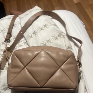 Beige quiltad axelväska i skinnimitation - Säljer en beige axelväska med quiltad design och justerbart axelband. Väskan har en mjuk, lyxig känsla och stängs med dragkedja. Perfekt storlek för det viktigaste och passar till många outfits.