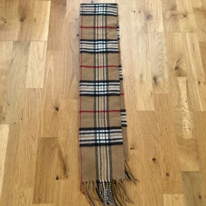 Rutig halsduk i ull  - Klassisk halsduk  i beige med rutigt mönster i svart, vitt och rött. Halsduken är lång med fransar i ändarna och tillverkad i mjuk ull. Perfekt accessoar för att lyfta din outfit med en tidlös brittisk vibe. Detta är inte Burberry men ser exaxr ut som en riktig. Som vanligt går priset alltid att ändra. 