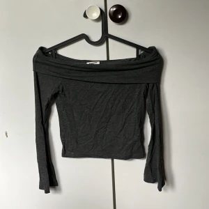 Mörkgrå offshoulder topp Kappahl  - Mörkgrå offshoulder topp från Kappahl med långa ärmar. Toppens breda kant upptill ger en snygg och avslappnad look. Mjuk och stretchig, perfekt att styla med jeans eller kjol för en trendig vibe.