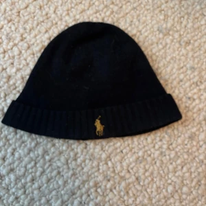 Svart mössa från Polo Ralph Lauren - Svart mössa från Polo Ralph Lauren i mjuk merinoull. Klassisk ribbad kant och ikonisk gul broderad logga framtill. Perfekt för kalla dagar och enkel att matcha med alla outfits.