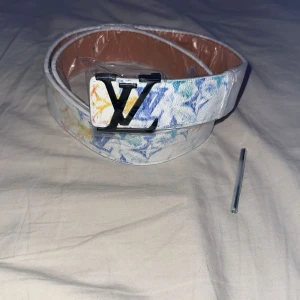 Vit bälte med färgglatt mönster - Snyggt vitt bälte med färgglada loggor i blått, gult och rött. Bältet har en stor svart -spänne Perfekt accessoar för att lyfta din outfit med en exklusiv touch.