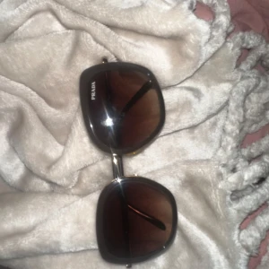 Bruna solglasögon från Prada - Snygga solglasögon från Prada med stora, fyrkantiga bågar i svart och bruna tonade glas. Klassisk och trendig design med diskret logga på sidan. Perfekta för att lyfta vilken outfit som helst. Pris kan diskuteras :)