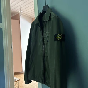 Grön overshirt från Stone Island - Säljer en grön overshirt från Stone Island med klassisk patch på vänster ärm. Skjortan har dragkedja framtill, bröstficka med knapp och en stilren krage. Perfekt för lager-på-lager och streetwear-stil. Storlek small men passar även medium skulle jag säga. 
