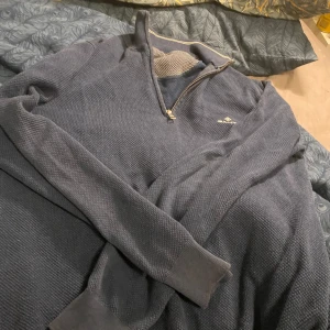 Blå halvzip tröja från GANT - Säljer en blå långärmad tröja från GANT med halv dragkedja och diskret logga på bröstet. Tröjan är stickad i ett strukturerat mönster och har ribbade muddar vid ärmslut och krage. Perfekt för lager-på-lager-stil, liten defekt på armbågen som syns på sista bilden men är absolut inget man tänker på och borde vara väldigt enkelt att fixa.