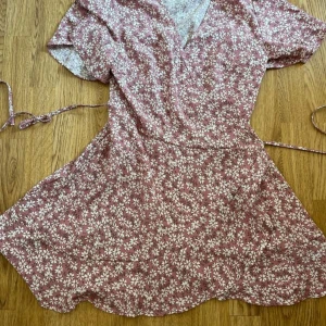 Rosa blommig omlottklänning från SHEIN - Söt rosa omlottklänning från SHEIN med vitt blommönster. Klänningen har korta ärmar, knyt i midjan och en lätt utställd kjol. Perfekt för varma dagar och ger en romantisk känsla till din stil.