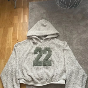 Grå teddy hoodie med siffran 22 - Mysig grå teddy hoodie med stor luva och grönt siffratryck '22' på bröstet. Croppad modell med breda muddar nertill. Perfekt för en avslappnad och trendig look.