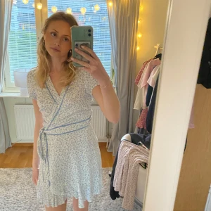 Ljusblå blommig omlottklänning - Söt ljusblå omlottklänning med små blommor och volangkant. Klänningen har korta ärmar, knyt i midjan och v-ringning. Perfekt för sommarens alla tillfällen. Materialet känns lätt och luftigt.