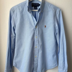 Ljusblå skjorta från Ralph Lauren - Klassisk ljusblå skjorta från Ralph Lauren med broderad logga på bröstet. Skjortan har lång ärm, knappar framtill och button-down krage. Tillverkad i 100% bomull för en skön och stilren känsla.