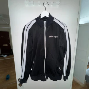 Svart track jacket från Palm Angels - Säljer en svart track jacket från Palm Angels med vita ränder längs ärmarna och dragkedja framtill. Jackan har hög krage, två fickor med dragkedja och logga på bröstet. Perfekt för en sportig och trendig look.