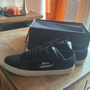 Svarta sneakers från Saint Laurent - Säljer ett par svarta sneakers från Saint Laurent med vit logga broderad på sidan. Skorna har svart snörning och vit platt sula. Perfekta för dig som gillar stilrena och exklusiva sneakers.