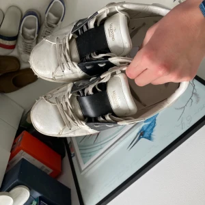 Valentino sneakers med svart detalj - Säljer ett par vita Valentino sneakers med bred svart rem över mitten och nitar på sulan. Skorna har rund tå och platt sula, samt klassisk snörning. Perfekta för dig som gillar exklusiva märken och stilren design.