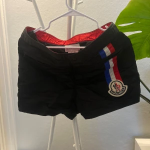 Moncler Badbyxor - Svarta shorts från Moncler med röd, vit och blå rand längs ena sidan samt Moncler-logga. Perfekta för sommaren. 