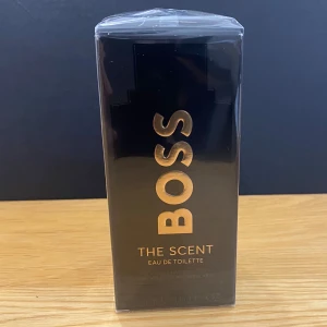BOSS The Scent Eau de Toilette herrparfym  - En elegant herrparfym från Hugo Boss, The Scent Eau de Toilette. Flaskan är cylindrisk med metalliska detaljer och bär en varm bärnstensfärgad vätska. Stilen är modern och lyxig, perfekt för dig som vill sticka ut med en sofistikerad doft.