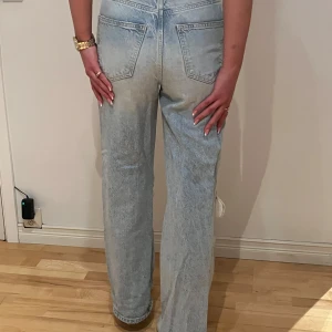 Ljusblå jeans med hål på knäna - Säljer ett par ljusblå jeans med raka ben och slitna hål på båda knäna. Jeansen har hög midja, klassiska fem fickor och stängs med dragkedja och knapp. Perfekta för en avslappnad och trendig look.