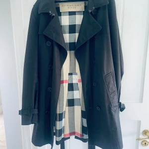 Svart trenchcoat från Burberry Brit - Klassisk svart trenchcoat från Burberry Brit med ikoniskt rutigt foder. Jackan har dubbelknäppning, bälte i midjan och vid ärmslut samt krage. Perfekt för dig som vill ha en tidlös och stilren jacka med lyxig känsla.