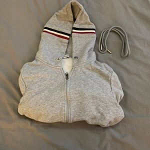  Moncler hoodie i grått – stilren och bekväm! - Säljer en snygg och stilren Moncler hoodie i ljusgrå färg med dragkedja. Klassisk design med röd, vit och marinblå detalj på huva och ärmslut, samt Monclers logotyp på ärmen.                                                            ✅ Väldigt bra skick – knappt använd ✅ Storlek: M men kan även passa S
