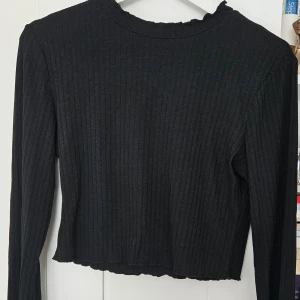 Svart ribbad croppad långärmad topp - Svart långärmad topp med ribbad struktur och croppad passform. Perfekt att matcha med höga jeans eller kjol för en trendig look.