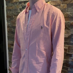 70% rabatt - Ralph Lauren skjorta  - Säljer en klassisk ljusrosa skjorta från Ralph Lauren med broderad logga på bröstet. Skjortan har knappar framtill och krage, perfekt för en stilren look. Passar dig som gillar tidlös stil. Skick: 10/10, perfekt. Använd en gång endast. Storlek: M. Kontakta vid intresse!