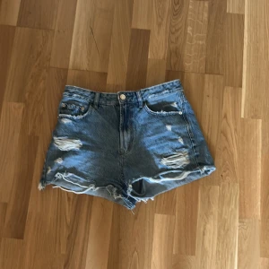 Blå jeansshorts från Stradivarius - Säljer ett par blå jeansshorts från Stradivarius med hög midja och slitna detaljer framtill. Shortsen har råa kanter nertill och klassiska fem fickor. Perfekta för sommaren!