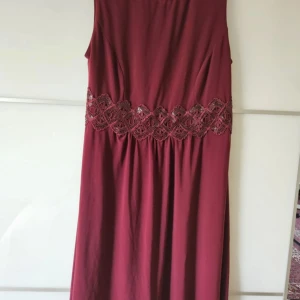 Vinröd ärmlös långklänning med spetsdetalj - Vinröd långklänning med vacker spetsdetalj i midjan. Klänningen är ärmlös och har en elegant, enkel design som passar perfekt till festliga tillfällen.