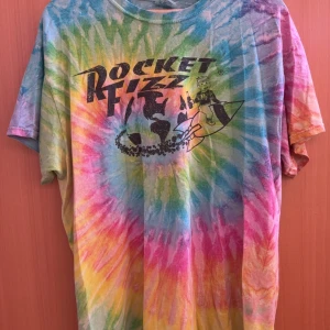 Färgglad tie-dye t-shirt från Colortone med Rocket Fizz-tryck - Säljer en riktigt färgstark tie-dye t-shirt från Colortone med stort svart Rocket Fizz-tryck på bröstet. T-shirten har klassisk passform och korta ärmar. Perfekt för dig som gillar unika och färgglada plagg!