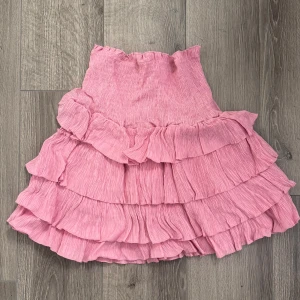 Rosa volangkjol med hög midja - Supersöt rosa kjol med flera lager volanger och hög midja. Kjolen har en kort längd och är perfekt för dig som gillar en feminin och lekfull stil. Materialet känns lätt och luftigt, vilket gör den skön att bära under varmare dagar.