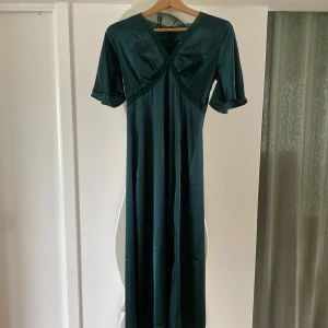 Grön långklänning med spetsdetaljer - Elegant långklänning i djupgrön nyans med v-ringning och spetsdetaljer längs halsringning, byst och slits. Klänningen har korta ärmar och är tillverkad i ett glansigt material som ger en lyxig känsla. Har aldrig använt den, prislapp sitter kvar. Säljer denna i mörkbrun oxå (se profil). 