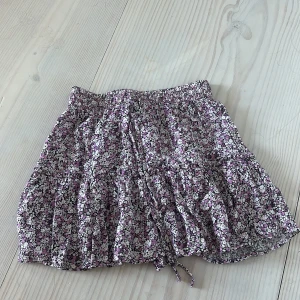 Blommig kortkjol med volang - Supersöt kortkjol med blommigt mönster i lila, rosa och vitt. Kjolen har volanger och resår i midjan samt knytband framtill. Perfekt för en somrig och avslappnad stil.