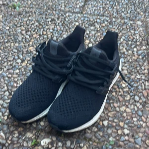 Svarta Adidas Ultraboost sneakers - Säljer ett par svarta Adidas Ultraboost sneakers med vit sula och svart ovandel i mesh. Skorna har snörning och klassiska tre ränder på sidan. Perfekta för träning eller vardagsstil.