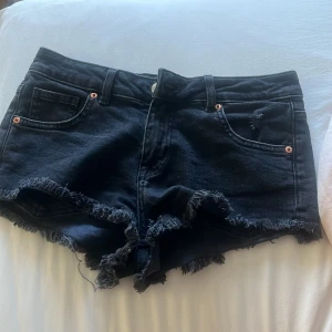 Svarta jeansshorts med fransar - Säljer ett par svarta jeansshorts med fransiga kanter och klassisk femficksdesign. Shortsen har låg midja och kopparfärgade nitar vid fickorna. Perfekta för varma dagar och enkel att matcha med olika toppar.( ståg inte storlek men skulle passa nån som har xs-s) 