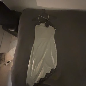 Satin slipklänning med spetsdetaljer - Säljer en ljusgrå lång slipklänning i satin med tunna axelband och svarta spetsdetaljer. Perfekt för dig som gillar en elegant och enkel stil. Klänningen har en mjuk glans och följsam passform. (Nyskick/oanvänd).